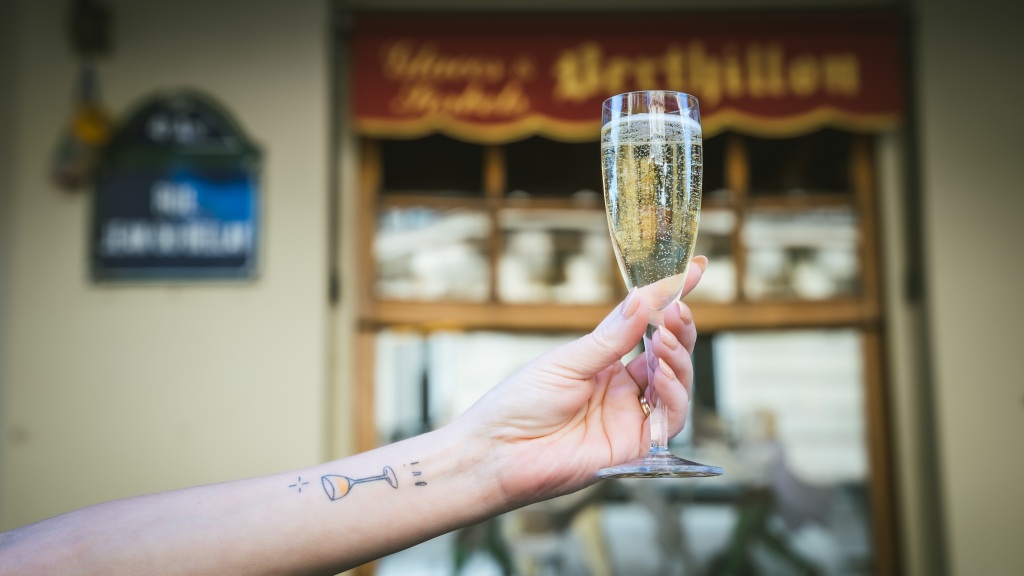 A hand holding a flute of champagne. The wrist is tattoed with a coupe of champagne and the world oui
